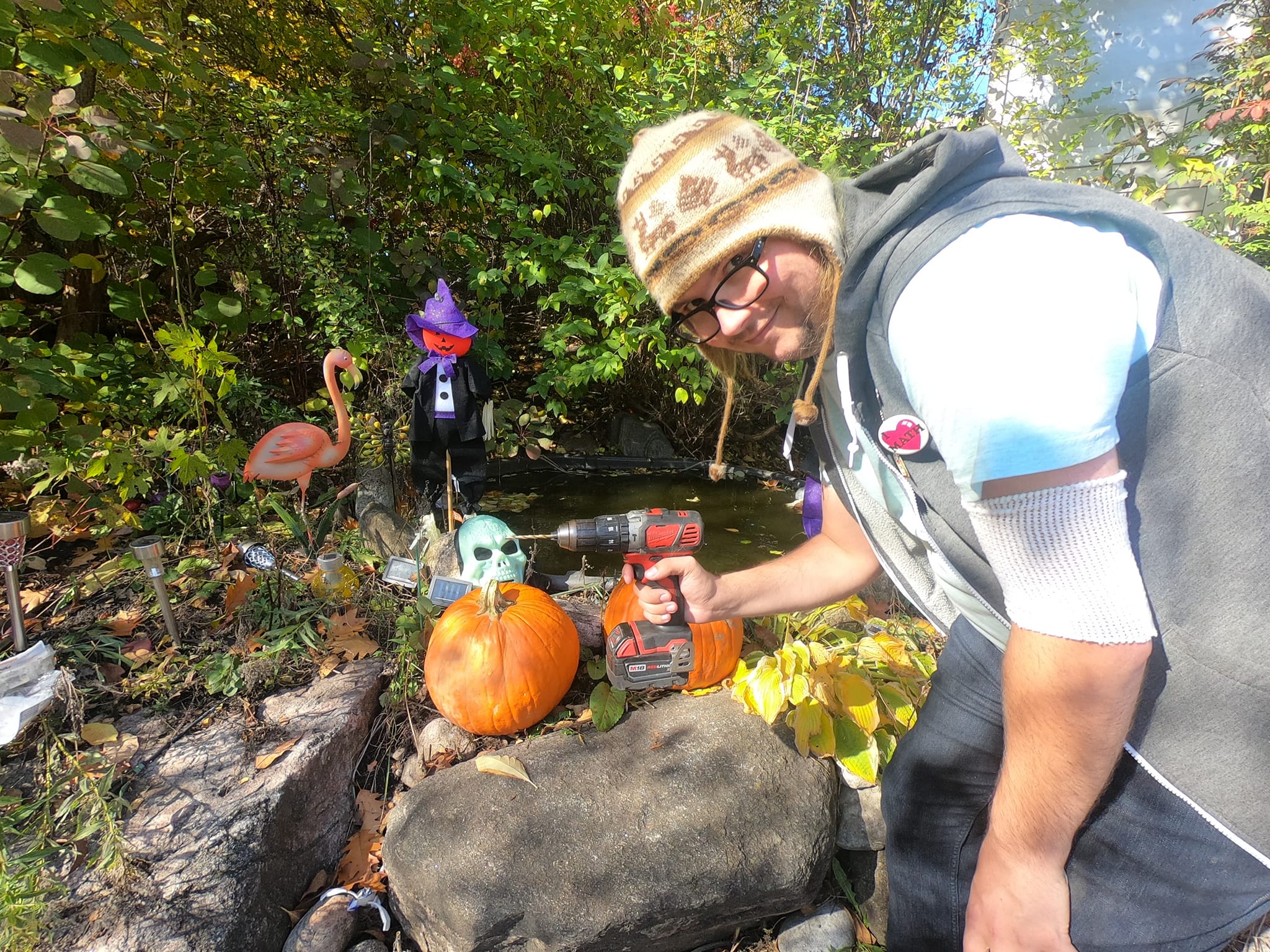 A man is wearing a toque, and is holding a cordless drill menacingly over two large pumpkins